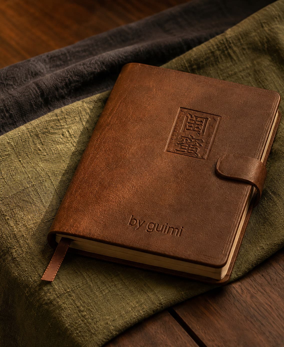 A brown leather notebook embossed with 闺蜜 and by guimi, resting on olive linen